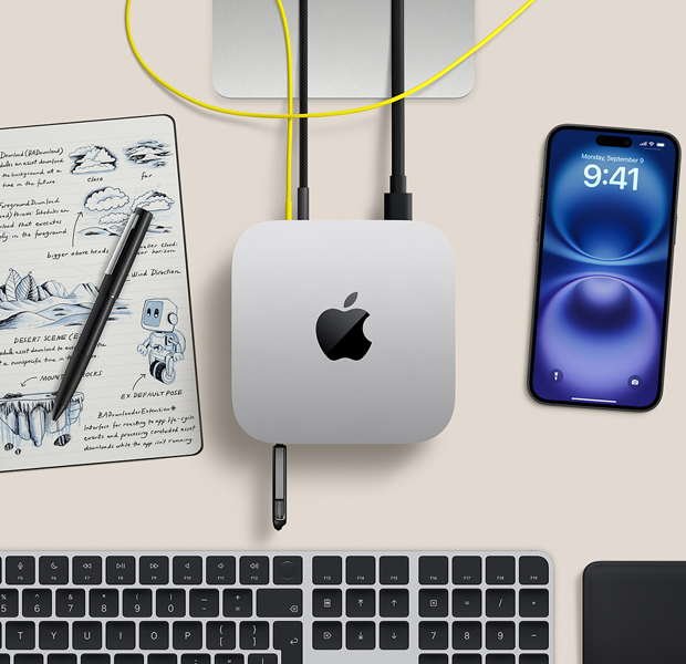 Imagem de cima do Mac mini prateado sobre uma mesa com um pen drive e cabos conectados às portas, ao lado de um teclado sem fio, trackpad, iPhone e bloco de notas.