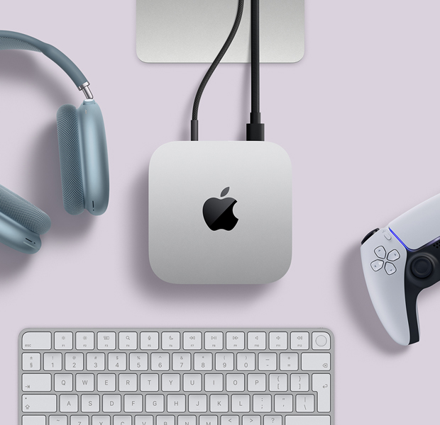 Imagem de cima do Mac mini prateado, com dois cabos conectados às portas de trás, ao lado de AirPods Max azuis, um Magic Keyboard prateado e um controle sem fio para jogos.