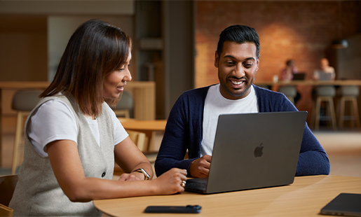 Amar collaborating with a colleague while sitting with his MacBook at a table in a common area.