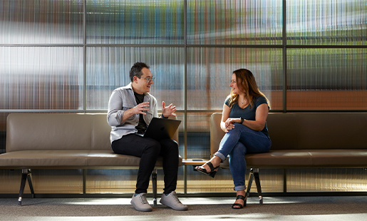 Dan sitting on an office bench with his MacBook on his lap and speaking with a colleague