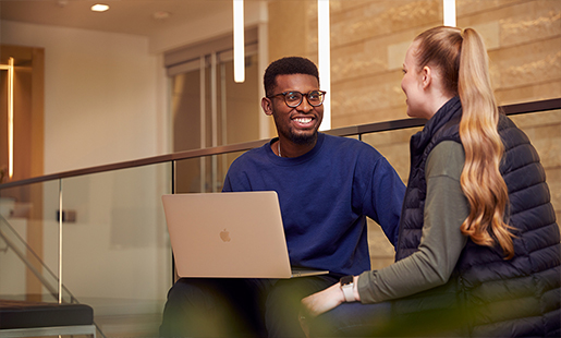 Chidi hat ein MacBook auf dem Schoß, während er mit einer Kollegin spricht. 