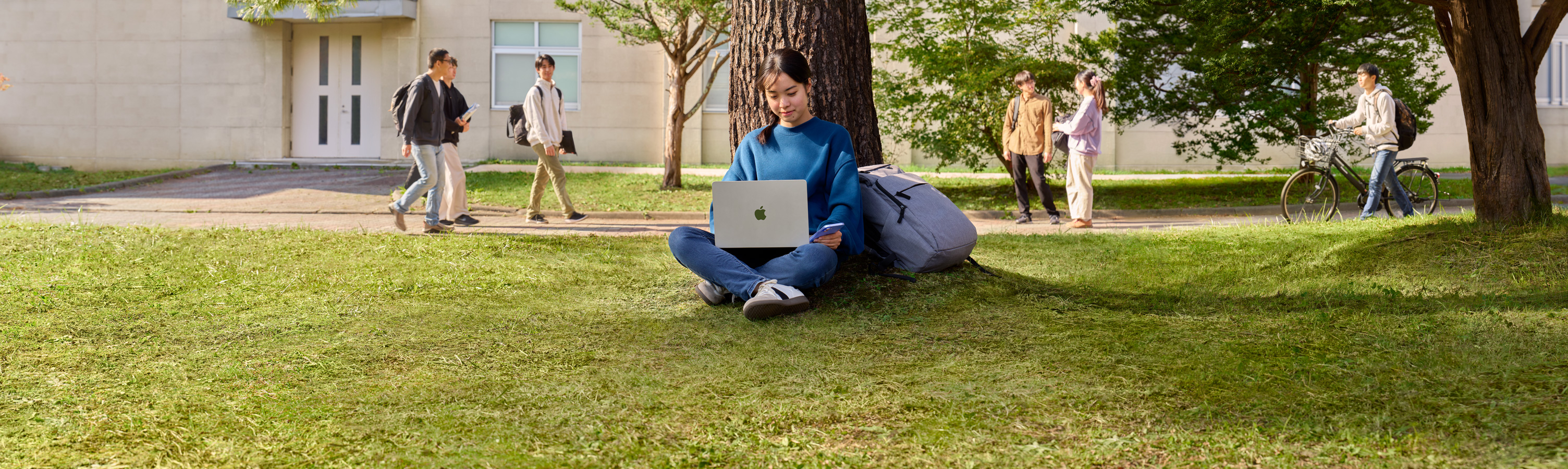 大学のキャンパスで大きな木の前に座っている学生がMacBook AirとiPhoneを使っている。