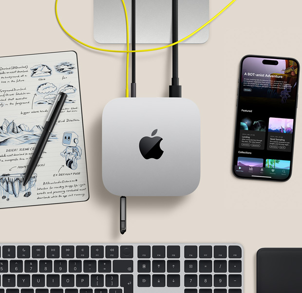 Top view of silver Mac mini with cords and a flash drive plugged into ports, in a desk setup with wireless keyboard, trackpad, iPhone, and notepad