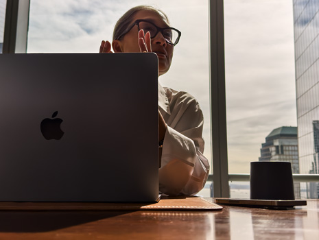 A woman sitting at a table in a high-rise building. Behind her is a large window revealing other buildings. In front of her is an open MacBook.