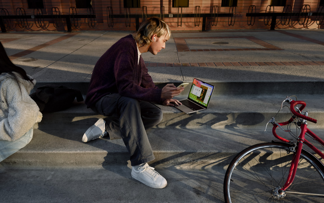 自転車のそばで階段に座ってiPhoneとMacBookを使っている学生