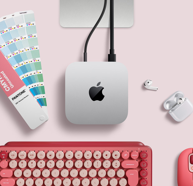 Top view of silver Mac mini in a desk setup with AirPods, a pink wireless keyboard, a pink wireless mouse and a colour swatch fan