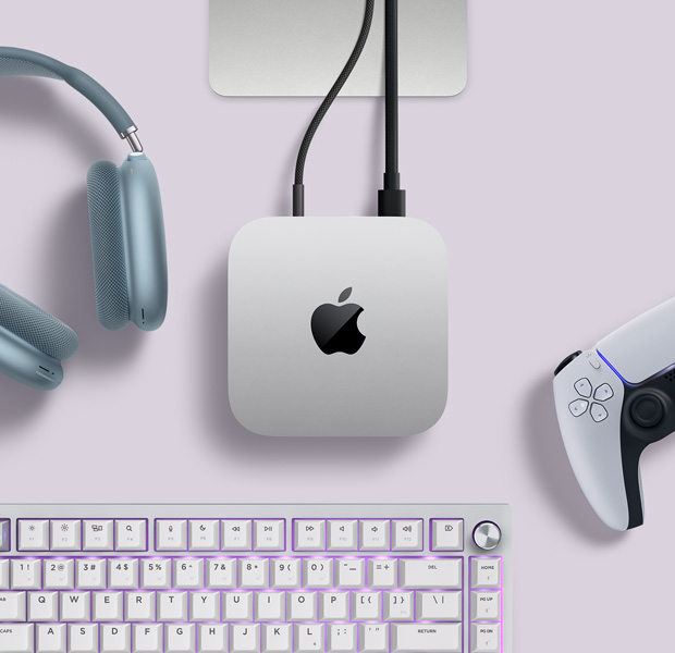 Top view of silver Mac mini, with two cords plugged into back ports, next to blue AirPods Max, a silver Magic Keyboard and a wireless game controller