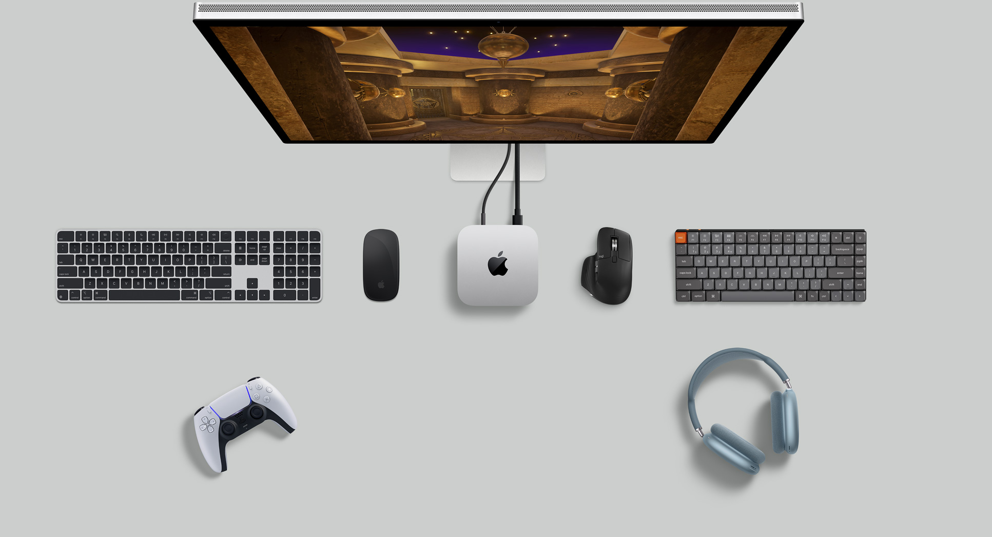 Top view of silver Mac mini in a desk setup with a display screen, wireless keyboards, wireless mice, a gaming controller and AirPods Max