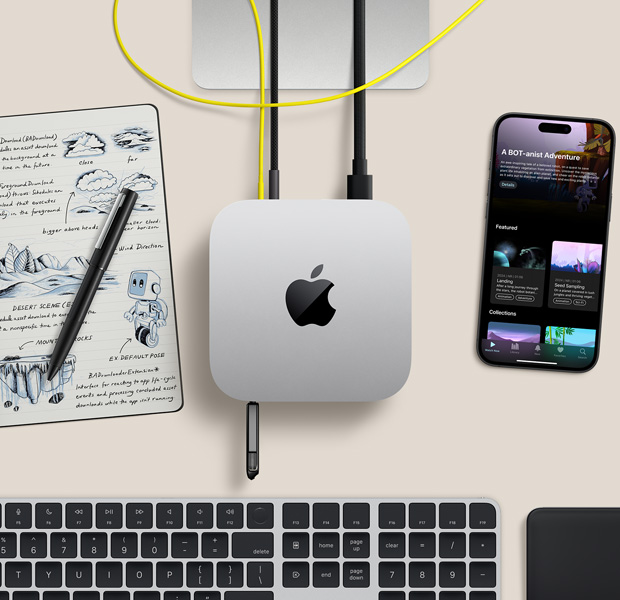 Top view of silver Mac mini with cords and a flash drive plugged into ports, in a desk setup with wireless keyboard, trackpad, iPhone and notepad